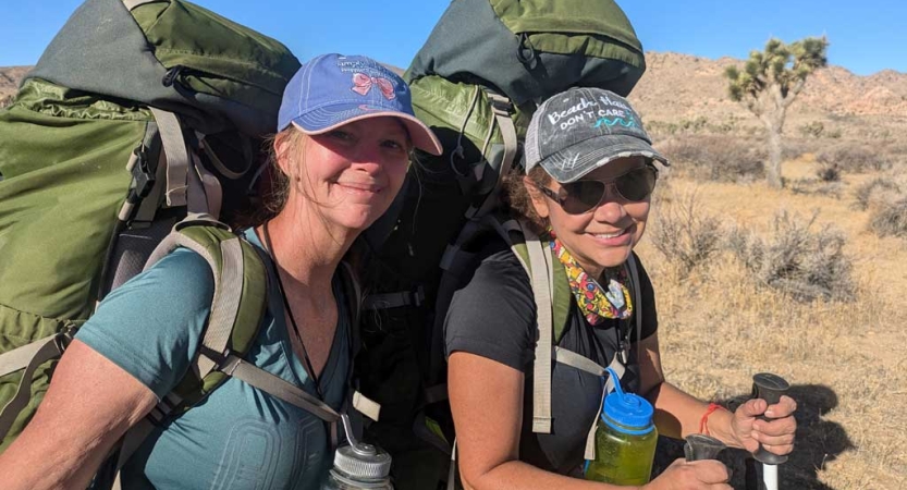 Two people wearing backpacks smile for the photo in a desert landscape. 